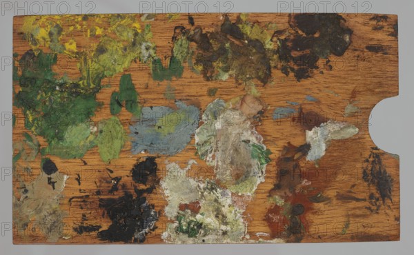 Jan Stanislawski's painting palette; Jan Stanislawski 1860–1907, painter; painting accessories; painting palettes; plein-air painting; painter's workshop; late 19th century; varia; height 8.7 cm, width 14.5 cm, A wooden palette with a warm brown grain visible across its surface, covered with a group of dried paint smears and blobs in several colors including greens, yellows, black, white, gray, bluish-gray, brown, reddish-brown, and small areas of pale beige; the paint is applied in thick textured patches and smudges across the palette, with larger green and yellow concentrations toward one side, darker black and brown areas in other spots, and mixed creamy white and gray masses near the center, all set against the wood background with a thumb cutout visible on one edge.