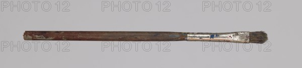 Jan Stanislawski's flat brush; Jan Stanislawski 1860–1907, painter. Painting accessory flat brush used in a painter's workshop and for plein-air painting, late 19th century. Dimensions: height 12.5 cm, width 0.5 cm., A single used paintbrush lies horizontally across a light gray background, with a long worn wooden handle showing reddish-brown and darker brown areas and spots of pale green paint, a metallic ferrule near one end that is scratched and mottled with silver and small splashes of blue and red, and a short cluster of dark bristles at the tip that appear stained and frayed.