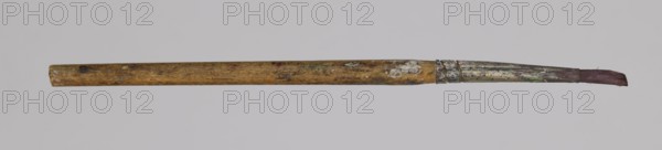 Jan Stanislawski's round brush; maker: Jan Stanislawski 1860–1907 painter; painting accessory: round brush for plein-air painting; late 19th century; dimensions: height 13.5 cm, width 0.7 cm; category: brushes, painting accessories, varia., A single used paintbrush lies horizontally against a pale gray background; the wooden handle is worn and mottled with stains in brown and yellowish tones, showing a few darker spots and areas of lighter discoloration, the metal ferrule is tarnished and splattered with remnants of dried paint in muted green, white, and silvery gray, and the bristles at the tip appear frayed and stained a dull reddish-brown.