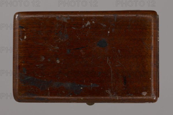 Jan Stanislawski's painting box; maker: Jan Stanislawski 1860–1907 painter; painting implements; artists in the open air; painting accessories; plein air studies; late 19th century; varia; dimensions: height 10 cm, width 16 cm, depth 3.5 cm, A rectangular wooden box with rounded corners seen from above, showing a dark brown polished surface with visible wood grain and a few scattered small dark spots and lighter scuffs, several scratches and worn areas, and a small metallic hinge or latch protruding slightly at one edge; the background is a neutral gray.