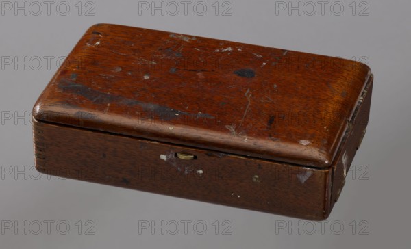 Jan Stanislawski's painting box; Jan Stanislawski 1860–1907, painter; painting implements; artists in the open air; painting accessories; plein air studies; late 19th century; varia; dimensions: height 10 cm, width 16 cm, depth 3.5 cm., A rectangular wooden box with rounded top edges sits against a neutral gray background; the box shows a worn brown finish with visible wood grain and several scuffed and chipped areas that reveal lighter and darker patches, along with a few small spots of darker staining and some pale scratches; a small metallic latch is visible on the front edge, and the right side shows a hinge with worn metal and a few pale marks; overall the colors present include brown, dark brown, black, gray, pale beige, and small touches of metallic gold.