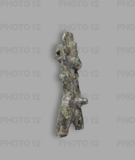 Mahes figurine, Mahes mythology. Uraeuses. Figurine of a walking ithyphallic male figure with a lion's head. On the head is a tripartite wig with a solar disk and a large uraeus. The left hand grasps the phallus; the right is raised upwards. A hanging ring is attached to the back. Roman Period, Egypt. Terracotta figurine. Height 6 cm, width 2.1 cm., A roughly vertical, irregular shaped object with a textured, pitted surface that appears metallic and weathered, showing patches of green, brown, dark gray, and hints of lighter gray; the object has a few protrusions and rounded nodules, an opening near the upper portion, and a split toward the lower end forming two leg-like extensions, all set against a plain light gray background.