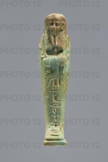 Ankhren's shabti, Ankh-ren name. Mummy-shaped figurine, funerary shabti, standing on a base and supported at the back by a pilaster. Arms crossed on the chest, holding hoes in both hands; in the right hand also a string from which a basket is slung over the left shoulder. Convex tools. Smooth three-part wig, ears uncovered. Facial details slightly blurred, large eyes, proportionate nose, straight long beard. Inscription engraved in a column on the front of the legs: The word spoken by Osiris Ankh-ren .... Date: 664–30 BC Late Period–Ptolemaic Period. Provenance: Tuna el-Gebel. Height: 10.8 cm., A small human-shaped figurine with a rounded head and stylized facial features, its arms crossed over the chest and the body tapering down to a narrow base, showing a worn surface with small pits and irregularities; the object displays a patina of green and turquoise with areas of brown and tan, set against a plain neutral gray background.