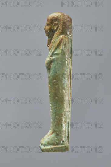 Ankhren's shabti, Ankh-ren name. Mummy-shaped figurine shabti, funerary gift, standing on a base and supported at the back by a pilaster. Arms crossed on the chest, holding hoes in both hands; a string in the right hand suspends a basket over the left shoulder. Convex tools. Smooth three-part wig, ears uncovered. Facial details slightly blurred, large eyes, proportionate nose, straight long beard. Inscription engraved in a column on the front of the legs: The word spoken by Osiris Ankh-ren .... Date: 664–30 BC Late Period–Ptolemaic Period. Provenance: Tuna el-Gebel Tuna el-Galat according to owner. Type: shabti. Height: 10.8 cm., A small standing figure seen in profile against a plain gray background, with a smooth elongated shape and a stylized head and shoulders; the surface shows a mottled green glaze with areas of darker green, brown, and pale tan, and a worn, slightly glossy texture; the figure appears wrapped or draped with subtle raised contours suggesting arms and a narrow base...