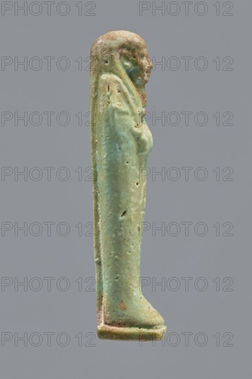 Ankhren's shabti, Ankh-ren name. Mummy-shaped figurine, funerary shabti, standing on a base and supported at the back by a pilaster. Arms crossed on the chest, holding hoes in both hands; in the right hand also a string suspending a basket over the left shoulder. Convex tools. Smooth three-part wig, ears uncovered, facial details slightly blurred with large eyes, proportionate nose, straight long beard. Inscription engraved in a column on the front of the legs: The word spoken by Osiris Ankh-ren .... Date: 664–30 BC; Late Period–Ptolemaic Period. Provenance: Tuna el-Gebel. Type: shabti. Height: 10.8 cm., A small vertical statuette of a standing human figure shown in profile against a neutral gray background, primarily green with areas of lighter green and pale turquoise patina and some brownish and tan weathered spots, with a smooth elongated form, a defined head and facial profile, a long cloak or garment that drapes down the back, subtle indications of arms and a torso, surface pitti...