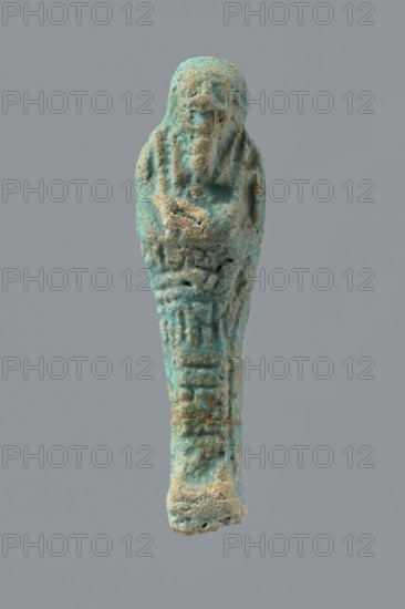 Ushabti, mummy-shaped funerary figurine wrapped in a shroud with its arms crossed on its chest, holding hoes. Inscription on the front in the form of the letter T. 332–30 BC; Ptolemaic Period, Egypt. Ushabti; height 10 cm, width 2.6 cm., A small weathered figurine shaped like a wrapped human form with a rounded head and tapered body, showing a textured surface with rough patches and erosion; the visible colors are turquoise blue, pale green, sandy beige, and spots of darker brown, set against a plain gray background.