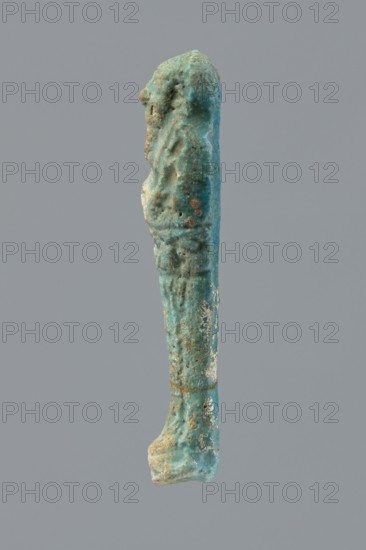 Ushabti, Mummy-shaped figurine funerary offering, wrapped in a shroud with its arms crossed on its chest, holding hoes. Inscription on the front in the form of the letter T. 332–30 BC; Ptolemaic Period, Egypt. Ushabti; height 10 cm, width 2.6 cm., A vertical elongated object against a plain gray background that is predominantly turquoise with areas of darker teal and lighter aqua, showing rough, pitted and irregular texture with patches of white and small rusty brown spots, an indented upper section with subtle raised forms along its length, a slightly wider base that appears rounded and worn, and an overall uneven, weathered surface.
