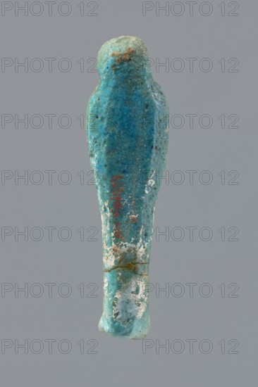 Ushabti, mummy-shaped funerary figurine wrapped in a shroud with its arms crossed on its chest, holding hoes. Inscription on the front in the form of the letter T. 332–30 BC; Ptolemaic Period, Egypt. Ushabti, height 10 cm, width 2.6 cm., A small elongated figurine-like object with a rounded top and tapering body, shown against a plain gray background; the object's surface is a mix of turquoise and blue with areas of lighter greenish-blue and pale white encrustation, and a few rusty brown spots and darker speckles scattered across its textured surface.