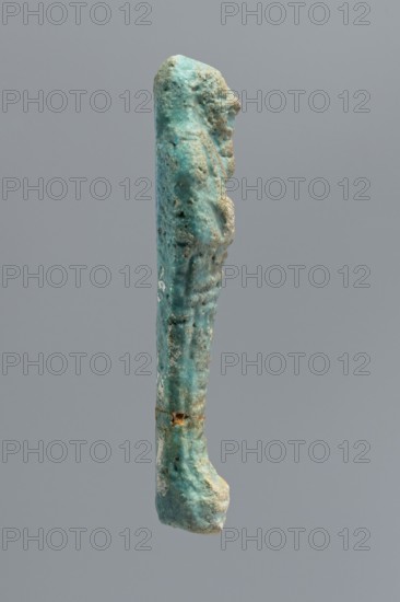 Ushabti, mummy-shaped funerary figurine wrapped in a shroud with its arms crossed on its chest, holding hoes. Inscription on the front in the form of the letter T. 332–30 BC; Ptolemaic Period, Egypt. Ushabti; height 10 cm, width 2.6 cm., A narrow vertical object with a rough textured surface predominantly in shades of turquoise and aqua, showing areas of pale blue-green and spots of sandy beige and light brown; the object has irregular contours with a rounded top and a thicker, slightly bulbous lower end, and a few small pitted areas and crusty mineral-like deposits along its length, all set against a uniform gray background.