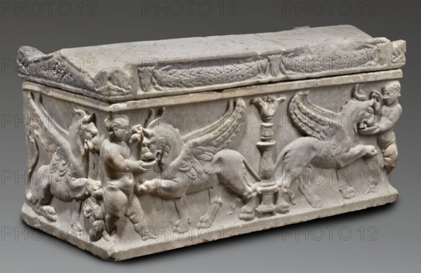 Children's sarcophagus with a depiction of griffins, The lid is shaped like a roof with tympanums; the longer frontal side is decorated with two garlands attached to three bucranions placed symmetrically at the sides and in the middle. On the shorter sides, between the tympanums, are double rosettes with ribbons. In the center of the front wall is a candelabrum topped with a head in the shape of a leaf. On either side of the candelabrum are symmetrically arranged winged griffins with lion heads, additionally decorated with ram's horns; both fantastical animals drink from goblets handed to them by naked boys placed symmetrically at the edges of the sarcophagus. The boys pour an unidentified drink from jugs held in their right or left hands. The side walls depict griffins in a sitting position with one paw resting on a recumbent ram's head. Iconographically, the griffins follow types traceable to Greek archaic art, reappearing on Roman sarcophagi and urns as guardians ensuring the peace ...