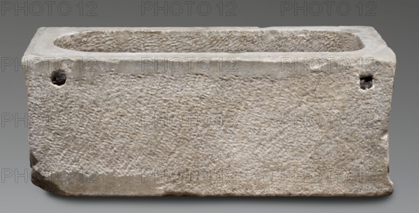 Children's sarcophagus with a depiction of griffins, Lid fashioned like a roof with tympanums; longer frontal side decorated with two garlands attached to three bucrania placed symmetrically and an additional bucranium in the lid's middle. Shorter sides show double rosettes with ribbons between tympanums. Center of the front wall features a candelabrum topped with a leaf-shaped head. On either side of the candelabrum are symmetrically arranged winged griffins with lion heads and ram's horns, drinking from goblets offered by naked boys at the sarcophagus edges; the boys pour a drink from jugs held in their right or left hands. Side walls depict seated griffins similar to those on the front, each with one paw on a recumbent ram's head. Iconography traces griffin types to Greek archaic art; they reappear on Roman sarcophagi as guardians ensuring the peace of the deceased. Hadrianic period c. 117–138 AD. Roman Empire. Osteotheca, sarcophagus. Dimensions: height 33 cm, width 68 cm, depth 29...