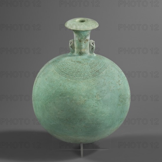 New Year's canteen. Flat round faience canteen with a narrow short neck ending in a chipped spout shaped like a papyrus capital. Both sides decorated with modeled figurines of crouching baboons elbows on knees, paws supporting the muzzle. A floral necklace in relief decorates both sides of the neck, and a hieroglyphic text runs along the narrow sides. Materials: faience. Function: vessel. Dimensions: height 17.5 cm +1.5 cm base, width 13.7 cm, weight 0.37 kg. Date: 664–525 BC, Late Period, 26th Dynasty, Egypt. Makers: unknown., A round, flattened vessel with a narrow flared neck and a wide rim sits against a plain gray background, the vessel showing a greenish-blue patina with areas of darker green and subtle brown spots; a decorative band of incised geometric patterns arcs below the neck, and two small sculpted figures flank the neck where it meets the body; the object rests on a clear support with a small vertical element visible at the base.