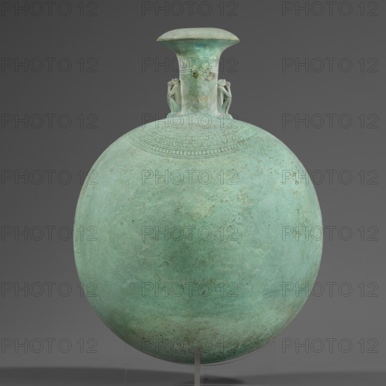 New Year's canteen, Goluchów Greater Poland Voivodeship. Flat round faience canteen with a narrow short neck ending in a chipped spout shaped like a papyrus capital. Decorated on both sides with modeled figurines of crouching baboons, elbows on knees and paws supporting the muzzle. A floral necklace is depicted in relief on both sides of the neck, and a hieroglyphic text runs along the narrow sides. Materials: faience. Function: ritual vessel/bottle. Date: 664–525 BC; Late Period, 26th Dynasty, Egypt. Height 17.5 cm +1.5 cm base; width 13.7 cm; weight 0.37 kg., A round vessel with a flattened spherical body and a short flaring neck set against a plain dark gray background, the vessel displaying a weathered greenish-blue patina with areas of darker green and small brownish spots; a band of incised geometric patterns decorates the upper shoulder just below the neck, and a pair of small sculpted humanlike figures flank the neck where it meets the body; the vessel rests on a clear stand th...