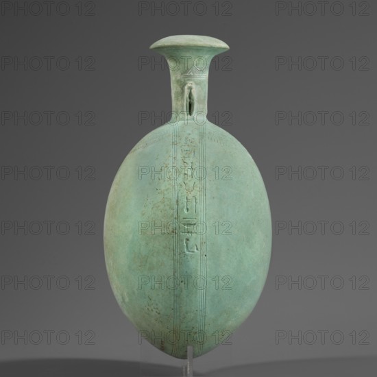 New Year's canteen, Flat round faience canteen with a narrow short neck ending in a chipped spout shaped like a papyrus capital, decorated on both sides with modeled figurines of crouching baboons elbows on knees, paws supporting the muzzle. A floral necklace is depicted in relief on both sides of the neck, and a hieroglyphic text runs along the narrow sides. Materials: faience. Function: vessel. Date: 664–525 BC, Late Period, 26th Dynasty, Egypt. Height 17.5 cm + 1.5 cm base, width 13.7 cm, weight 0.37 kg., A smooth, egg-shaped vessel in a pale green color stands upright against a neutral gray background, with a flared neck and lip in a slightly darker green; the body shows a vertical band of shallow incised lines running from neck to base and a faint pattern of small dotted spots and subtle discoloration in greens and brownish tones, and a small projection near the neck where the band meets the rim.