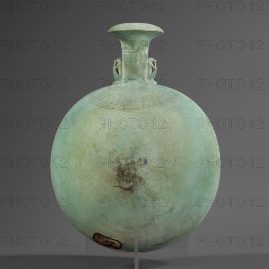 New Year's canteen, Flat round faience canteen with a narrow short neck ending in a chipped spout shaped like a papyrus capital, decorated on both sides with plastic figurines of crouching baboons with elbows on knees and paws supporting the muzzle. A floral necklace is depicted in relief on both sides of the neck, and a hieroglyphic text runs along the narrow sides. Date: 664–525 BC, Late Period, 26th Dynasty, Egypt. Vessel; height 17.5 cm +1.5 cm base, width 13.7 cm, weight 0.37 kg., A round, flattened vessel with a short flared neck and wide rim, shown upright on a clear stand against a plain dark gray background; the vessel surface is predominantly pale green with areas of light blue-green and subtle beige patina, and there is a darker irregular brownish stain near the center of its face; near the neck there is a band of incised decorative lines and geometric patterns, and two small applied sculptural elements at the shoulder on either side of the neck; a small fragment of a dark b...