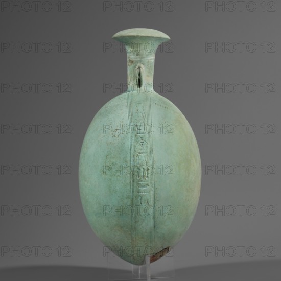 New Year's canteen, Goluchów Greater Poland Voivodeship. Flat round faience canteen with a narrow short neck ending in a chipped spout shaped like a papyrus capital, decorated on both sides with plastic figurines of crouching baboons elbows on knees, paws supporting the muzzle. A floral necklace is depicted in relief on both sides of the neck, and a hieroglyphic text runs along the narrow sides. Materials: faience. Function: vessel. Date: 664–525 BC, Late Period, 26th Dynasty, Egypt. Height 17.5 cm +1.5 cm base, width 13.7 cm, weight 0.37 kg., A smooth oval vessel with a narrow neck and flared rim standing upright against a plain gray background, the vessel showing a predominantly greenish-blue patina with areas of lighter green and faint brownish spots; a vertical raised band runs along the front of the body with shallow incised shapes, and a small slit-like opening appears near the base of the neck; the vessel rests on a transparent stand that is partially visible and the overall lig...