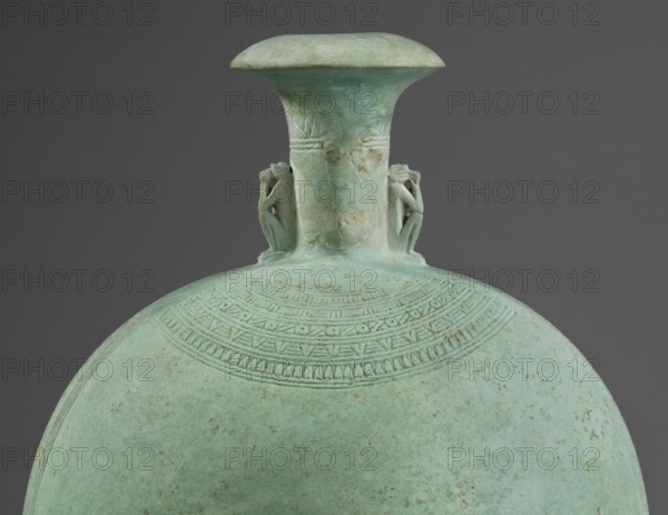 Flat round faience canteen with a narrow short neck ending in a chipped spout shaped like a papyrus capital, decorated on both sides with plastic figurines of crouching baboons with elbows on knees and paws supporting the muzzle. A floral necklace is depicted in relief on both sides of the neck, and a hieroglyphic text runs along the narrow sides. Height 17.5 cm plus 1.5 cm base; width 13.7 cm; weight 0.37 kg. Dated 664–525 BC, Late Period, 26th Dynasty, Egypt., A turquoise-green rounded vessel with a narrow neck and flared rim is shown against a dark gray background; the vessel surface has a patinated, slightly mottled texture with areas of lighter and darker green and small brownish spots, and a band of incised decorative geometric patterns encircles the upper shoulder; a pair of small sculpted human figures in the same green material are attached on opposite sides of the neck, each seated and holding their knees.