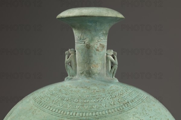 Flat round faience canteen with a narrow short neck ending in a chipped spout shaped like a papyrus capital, decorated on both sides with plastic figurines of crouching baboons with elbows on knees and paws supporting the muzzle. A floral necklace is depicted in relief on both sides of the neck, and a hieroglyphic text runs along the narrow sides. Height 17.5 cm plus 1.5 cm base; width 13.7 cm; weight 0.37 kg. Dated 664–525 BC, Late Period, 26th Dynasty, Egypt., A greenish-blue vessel with a tall flared neck and a rounded body occupies the image against a dark gray background; the vessel surface shows a weathered patina with areas of lighter and darker green-blue and some brownish spots, and a group of small sculpted figures in relief cling to the neck on opposite sides, while the rounded body is decorated with several concentric bands of incised geometric patterns and textured lines in the same green-blue tones.