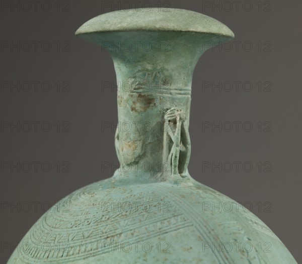 Flat round New Year’s canteen with a narrow short neck ending in a chipped spout shaped like a papyrus capital, decorated on both sides with plastic figurines of crouching baboons elbows on knees, paws supporting the muzzle. A floral necklace is depicted in relief on both sides of the neck, and a hieroglyphic text runs along the narrow sides. Faience vessel, 664–525 BC; Late Period, 26th Dynasty, Egypt. Height 17.5 cm plus 1.5 cm base, width 13.7 cm, weight 0.37 kg., A close-up of the upper portion of a rounded vessel with a flared neck and wide rim, showing a weathered surface in shades of green and turquoise with patches of pale brown and darker green patina, subtle speckling, and areas of erosion. A small sculpted attachment resembling a stylized human or figurative motif is visible at the base of the neck in the same green-turquoise material. The shoulder of the vessel displays concentric incised decorative bands and linear patterns following the curve, rendered in the same green a...