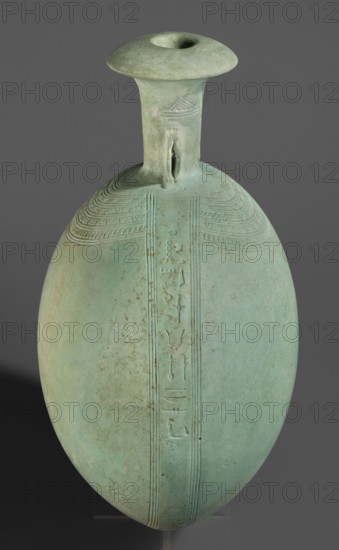 New Year's canteen, Flat round faience canteen with a narrow short neck ending in a chipped spout shaped like a papyrus capital, decorated on both sides with plastic figurines of crouching baboons with elbows on knees and paws supporting the muzzle. A floral necklace is depicted in relief on both sides of the neck, and a hieroglyphic text runs along the narrow sides. Material: faience. Function: vessel. Date: 664–525 BC; Late Period, 26th Dynasty, Egypt. Height 17.5 cm + 1.5 cm base, width 13.7 cm, weight 0.37 kg., A single elongated oval vessel with a narrow neck and flared rim, rendered in a greenish-blue patina with areas of lighter green and pale turquoise, sits against a neutral gray background; the surface shows a few scattered brownish spots and subtle weathering. The neck has a small applied loop handle and a raised decorative band near its base, and a vertical raised strip runs down the front of the body, bordered by several fine incised parallel lines and bearing a series of ...