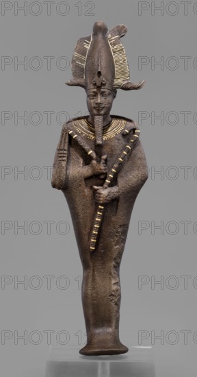 Figurine of the god Osiris; mummy-shaped male figurine; Egyptian art; Egyptian deities; crowns; Osiris myth.; sculpture;, Goluchów Greater Poland Voivodeship - place associated with object; 664–525 BC, Late Period, 26th Dynasty, Egypt; height 29 cm, width 6.8 cm, depth 4 cm., A standing statuette of a human figure in a dark brown material with a smooth, slightly textured surface, wearing a tall headdress that has flared, ridged elements highlighted with gold accents, a long narrow beard, and a broad collar with gold decorative lines; the figure’s arms are crossed over its chest holding two long striped staffs with alternating dark and gold bands, the body tapers down to a rounded base, and the background is a uniform gray.