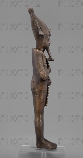Figurine of the god Osiris; mummy-shaped male deity sculpture, painted; Egyptian, Late Period, 26th Dynasty, 664–525 BC; height 29 cm, width 6.8 cm, depth 4 cm; inscriptions/attributes of Osiris crowns, iconography., A bronze statuette of a standing figure shown in profile on a clear rectangular base, the figure facing right with a tall conical headdress and a long braided beard; the surface shows a mottled dark brown and black patina with areas of greenish and golden highlights, and a few spots of lighter brown corrosion; the figure's arms are held close to the body, hands grasping a long narrow object that has alternating dark and golden bands; the body is slender with smooth contours, visible textured pitting on the torso and legs, and the base of the feet resting flat on the clear support against a neutral gray background.