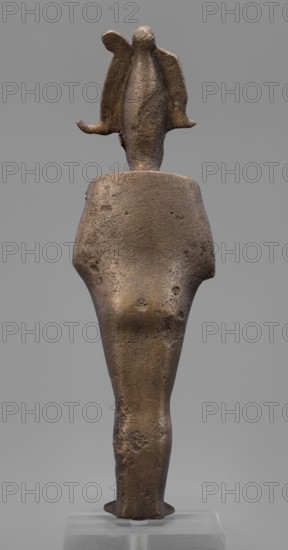 Figurine of the god Osiris;, Goluchów Greater Poland Voivodeship; Egyptian deities; crowns; mummy-shaped figurines; male figurines; Egyptian art; Osiris myth.; 664–525 BC, Late Period, 26th Dynasty, Egypt; sculpture; height 29 cm, width 6.8 cm, depth 4 cm, A single standing statuette viewed from the back, made of a rough textured material appearing brown and dark bronze with patches of lighter brown and small darker pitted spots; the figure has broad shoulders tapering to a narrow lower body and legs, with a rounded head topped by a winglike or petal-shaped element extending outward on each side; the surface shows uneven pits and abrasion, and the object stands on a clear rectangular base that appears translucent and pale gray, set against a neutral mid-gray background.