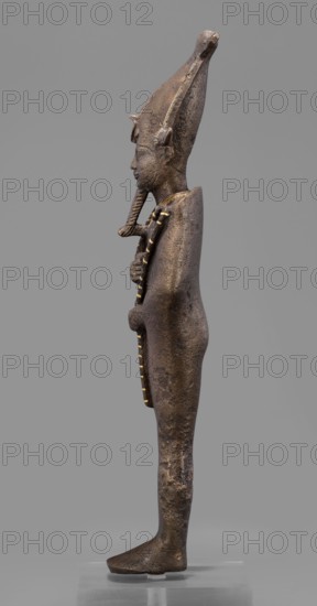 Figurine of the god Osiris;, Goluchów Greater Poland Voivodeship provenance; Egyptian deities; crowns; mummy-shaped figurines; male figurines; Egyptian art; Osiris myth.; 664–525 BC, Late Period, 26th Dynasty, Egypt; sculpture; height 29 cm, width 6.8 cm, depth 4 cm., A side view of a standing statuette in profile showing a slender human figure with an elongated conical headdress and a false beard; the surface of the figure is dark brown with areas of lighter brown and some subtle golden accents along the beard and vertical adornment on the chest and front. The figure stands with arms close to the body, one hand holding a staff-like object that runs down the torso, and the body and limbs show a textured, slightly pitted surface. The headdress has small projecting elements at the sides and the facial features are rendered in relief with a straight nose and closed lips. The base beneath the figure is a translucent support and the background is a uniform mid gray.