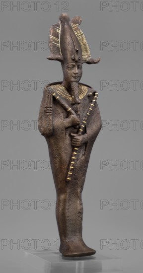 Figurine of the god Osiris; maker, Goluchów Greater Poland Voivodeship; Egyptian deities; crowns; mummy-shaped figurines; male figurines; Egyptian art; Osiris myth.; 664–525 BC, Late Period, 26th Dynasty, Egypt; sculpture; height 29 cm, width 6.8 cm, depth 4 cm., A standing statue of a human figure made of dark brown material with a textured surface, wearing a tall, ornate headdress with decorative ridged elements highlighted in gold, a stylized beard, and a broad collar with gold accents; the figure holds two crossed objects in front of the chest, one of which has alternating gold and dark segments, and the other shows gold detailing; the arms are close to the body and the robe-like garment is smooth and dark brown; the statue stands on a clear rectangular base against a neutral gray background, and visible colors include dark brown, gold, and gray.