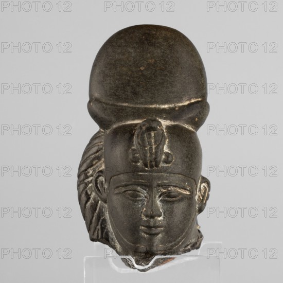Fragment of a figurine of the god Khonsu — head. Fragment of a figurine — head of the god Khonsu with a lock of youth and a disc mounted on a crescent moon on the head. A uraeus above the forehead; a false beard on the chin not preserved. Materials/format: figurine. Iconography: uraei, lunar discs and crescents, locks of youth, back pillars. Maker/attribution: unknown. Date: 664–525 BC; Late Period, 26th Dynasty, Egypt. Dimensions: height 7.4 cm, width 5 cm, depth 4 cm., A dark stone or metal sculpted head with a rounded tall headdress and a decorative element at the front, a smooth face with closed or relaxed eyes, a subtle smile, stylized ears, and detailed carved hair or headpiece elements on the sides, displayed against a light gray background with visible shades of black, dark brown, gray, and a few lighter worn highlights.
