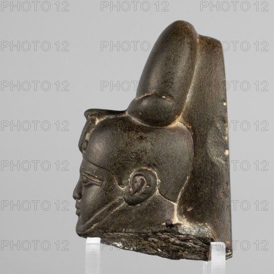Fragment of a figurine of the god Khonsu – head. Depicted wearing a lock of youth and a disc mounted on a crescent moon on the head; a uraeus above the forehead; a false beard on the chin not preserved. Egyptian, Late Period, 26th Dynasty, 664–525 BC. Material: figurine. Dimensions: height 7.4 cm, width 5 cm, depth 4 cm., A carved stone fragment showing a left-facing human profile with a pronounced ear, eyebrow ridge, nose, lips, and chin, topped by a tall rounded headdress that extends upward and slopes back; the surface appears dark brown to black with lighter brown highlights and some pale spots, and the lower edge shows rough, lighter-colored broken stone; the fragment rests on clear supports against a neutral light gray background.