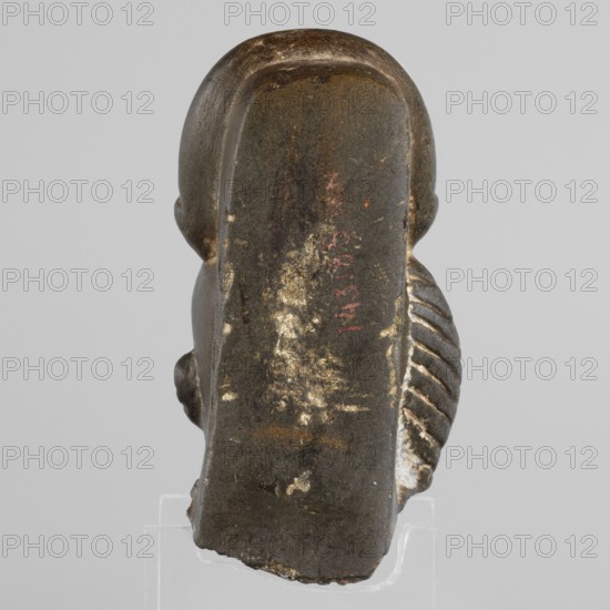 Fragment of a figurine of the god Khonsu — head with a lock of youth and a disc mounted on a crescent moon on the head; a uraeus above the forehead; a false beard on the chin not preserved. Materials and other identifiers: figurine, height 7.4 cm, width 5 cm, depth 4 cm. Date: 664–525 BC, Late Period, 26th Dynasty, Egypt. Subjects/icons: Khonsu mythology, uraei, lunar discs and crescents iconography, locks of youth, back pillars. Maker/attribution unknown., A small sculpted object shown from the back with a smooth rounded top and a flat vertical rear surface, predominantly dark gray to black with areas of worn lighter gray and off-white showing through, and a few small reddish marks; the sides reveal a rounded ear shape on one side and a series of carved parallel ridges on the other, both in similar dark tones with lighter worn highlights; the base is irregular and shows chipped edges with lighter interior color; the object rests on a clear display support against a pale gray backgroun...