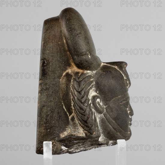 Fragment of a figurine of the god Khonsu — head with a lock of youth and a disc mounted on a crescent moon on the head; a uraeus above the forehead; a now-lost false beard on the chin. Egyptian, Late Period, 26th Dynasty, 664–525 BC. Material: figurine. Dimensions: height 7.4 cm, width 5 cm, depth 4 cm., A carved stone fragment showing the profile of a human head with a stylized headdress or crown, depicted in a dark gray to black stone with lighter worn highlights; the face is smooth with a defined nose, lips, ear, and an incised eye line, and the hair or headdress behind the ear is rendered with a braided or chevron pattern; the top of the fragment has a rounded protrusion and a curved crest, and the lower edge appears rough and broken; the object rests on two translucent stands against a neutral light gray background, and visible colors include dark gray, black, lighter gray highlights, and small areas of brownish discoloration.