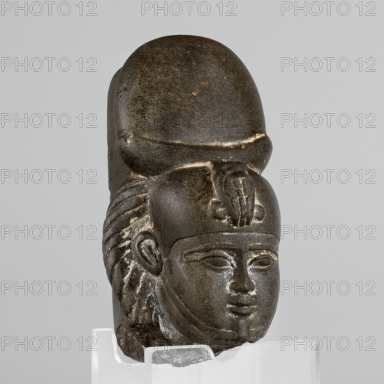 Fragment of a figurine of the god Khonsu — head. Fragment of a figurine: head of the god Khonsu with a lock of youth and a disc mounted on a crescent moon on the head; a uraeus above the forehead; a false beard on the chin not preserved., figurine. Date: 664–525 BC, Late Period, 26th Dynasty, Egypt. Dimensions: height 7.4 cm, width 5 cm, depth 4 cm., A carved stone head displayed on a clear stand, showing the front and part of the side of a stylized human face with almond-shaped eyes, a straight nose, and full lips; the figure wears a rounded headdress with a raised central emblem and a striped, textured wig or hair element at the side. The stone surface is dark gray to black with lighter worn areas and subtle brownish spots, and the clear stand is translucent white.