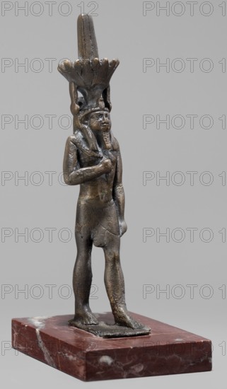 Figurine of the god Nefertum; creator, Egyptian deity Nefertum; Egyptian religion and art; crown depicted; Late Period, 664–332 BC; sculpture; height 12.4 cm, width 3.3 cm, depth 4 cm., A small bronze statuette of a standing human figure is shown on a rectangular reddish-brown stone base with lighter veining. The figure is dark bronze with areas of lighter patina and shows a person wearing a fitted short skirt and a tall ornate headdress that flares outward near the top. The figure stands with one arm bent across the chest and the other arm hanging down alongside the body, and the facial features include a stylized beard and a calm expression. The surface of the metal shows small irregularities and slight discoloration, and the background is a plain neutral gray.