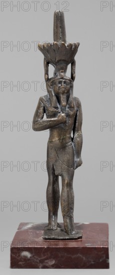 Figurine of the god Nefertum; creator, Egyptian deity Nefertum; Egyptian religion and art; crowns; Late Period, 664–332 BC, Egypt; sculpture; height 12.4 cm, width 3.3 cm, depth 4 cm., A small sculpted figure standing on a rectangular base, rendered in dark brown and bronze tones with areas of lighter brown and greenish patina; the figure wears a headdress with a tall vertical element and a flared, petal-like crown, both in similar dark metallic hues; the figure's facial features, beard, and hair are visible in the same metallic colors, and one arm is bent with the hand placed on the chest while the other arm hangs down; the torso and limbs show textured metal surface and subtle cracks or joins, and the base is a reddish-brown stone with white and lighter brown marbling.