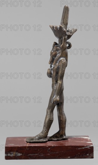 Figurine of the god Nefertum;, Egyptian deity Nefertum; Egyptian religion; Egyptian art; crowns; Late Period, 664–332 BC; Egypt; sculpture; height 12.4 cm, width 3.3 cm, depth 4 cm., A small metallic figurine in brownish bronze tones stands on a rectangular reddish-brown stone base with lighter veins; the figurine is shown in profile with a slender upright body, elongated legs, and flat feet, and it wears an elaborate headpiece that rises above the head and fans outward; the figure's torso and limbs show a slightly rough textured surface with darker and lighter bronze highlights, and a few smoother areas; the base appears polished and contrasts with the metallic figure; the background is a uniform light gray.