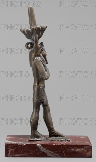 Figurine of the god Nefertum; creator, Egyptian deity Nefertum; Egyptian religion and art; crown; Late Period, 664–332 BC, Egypt; sculpture; height 12.4 cm, width 3.3 cm, depth 4 cm., A small metal statuette of a walking human figure stands on a rectangular reddish-brown stone base; the figure is shown in profile facing right with arms held close to the chest and wearing a tall decorative headdress that rises above the head and has a flared element and a looped detail at the back. The figure has a dark metallic surface with areas of lighter and slightly corroded texture, and the stone base shows veins and patches of lighter and darker red and gray. The background is a uniform light gray.