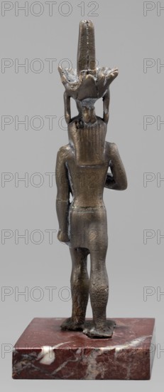 Figurine of the god Nefertum; Egyptian deity; sculpture in Egyptian art; material/creator, crowns; Late Period, 664–332 BC, Egypt; height 12.4 cm, width 3.3 cm, depth 4 cm., A small bronze figurine viewed from the back standing on a square reddish-brown marble base with white and lighter brown veins; the figure has a dark metallic bronze surface with areas of lighter patina and slight greenish corrosion, a tall central vertical headdress rising above a flared crown-like element, long straight hair or a backpiece reaching the shoulders, a fitted short garment around the waist, visible arms with one bent and the other hanging, and bare legs and feet positioned on the base; the background is a uniform light gray.