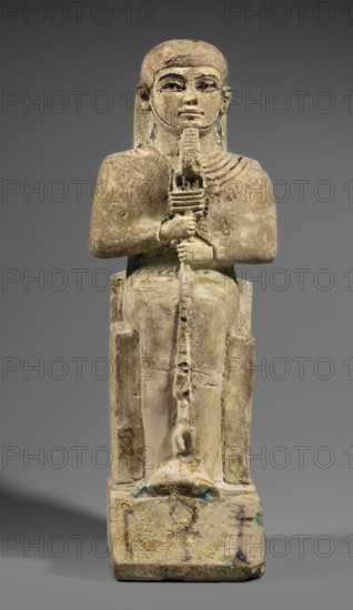 Figurine of the god Ptah sitting on a throne, on a base in the shape of the sign of Maat. He holds a composite scepter in his hands. Materials/attributes: ankh, uas, djed pillars, back pillars, hieroglyphs, scepters, thrones. Subject/style: Egyptian deity, figurine of a man. Maker/date: c. 1550–c. 1295 BC, New Kingdom, 18th Dynasty, Egypt. Dimensions: height 29 cm, width 10.5 cm, depth 19 cm, weight 2.736 kg., A seated stone figure with a rounded head and stylized facial features holds a long staff vertically with both hands; the figure wears a fitted garment and sits on a rectangular throne with a stepped base. The surface shows worn, textured stone with patches of beige, tan, and light brown, along with traces of darker brown and small areas of green-blue patina. The face and eyes are outlined in darker pigments, and there are subtle linear decorations across the chest in brownish tones. The background is a smooth gray.