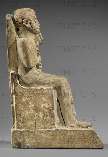 Figurine of the god Ptah sitting on a throne, on a base in the shape of the sign of Maat. He holds a composite scepter in his hands. Materials/attributes: hieroglyphs, ankh, uas, djed pillars, scepters, back pillars., Inscription/Historical details: c. 1550–c. 1295 BC; New Kingdom, 18th Dynasty, Egypt. Object type: figurine. Dimensions: height 29 cm; width 10.5 cm; depth 19 cm; weight 2.736 kg., A stone sculpture of a seated human figure viewed in profile, positioned on a rectangular block-like throne with a high straight back; the figure has a rounded head, a short stylized beard, and hands crossed and resting on the chest, with legs bent at the knees and feet extending forward to a sloped platform. The surface shows rough weathered texture and areas of wear, with overall beige and tan tones, along with patches of darker brown and faint greenish-blue discoloration in places. The background is a smooth neutral gray gradient.