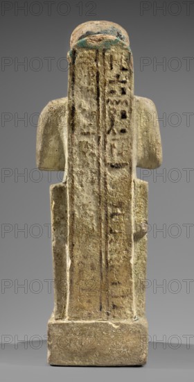 Figurine of the god Ptah. Figural sculpture of Ptah seated on a throne on a base in the shape of the sign of Maat, holding a composite scepter. Materials and stylistic elements: ankh, uas, djed pillars, back pillars, scepters, thrones; hieroglyphs; Egyptian deities iconography., Inscription references: Scen.Gal.Eg. Dated c. 1550–c. 1295 BC, New Kingdom, 18th Dynasty, Egypt. Figurine of men; height 29 cm, width 10.5 cm, depth 19 cm, weight 2.736 kg., A weathered vertical stone figurine viewed from the back, showing a rectangular base and elongated body with a flat back and slightly rounded shoulders; the surface has a rough, pitted texture with areas of erosion and faint linear incisions; visible colors include beige, sandy tan, light brown, dark brown accents, pale greenish-blue patches near the top, and subtle gray tones in the background; the figure's arms are low and close to the sides, and the top of the head is rounded with some darker discoloration.