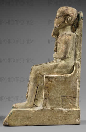Figurine of the god Ptah. Figurine of the god Ptah sitting on a throne, on a base in the shape of the sign of Maat. He holds a composite scepter in his hands. c. 1550–c. 1295 BC; New Kingdom, 18th Dynasty, Egypt. Materials/attributes: ankh, uas, djed pillars, back pillars, hieroglyphs, scepters, thrones, figurines of men., Height 29 cm, width 10.5 cm, depth 19 cm, weight 2.736 kg., A weathered seated figure carved in stone is shown in profile facing right, positioned on a blocky chair with a high back; the figure has a stylized beard and a hooked nose, with hands folded over the chest and legs extended forward so the feet rest on a low plinth. The surface shows rough, pitted texture and areas of erosion and small cracks, with traces of smoother carved detail around the face, ears, and necklace. The color palette consists mainly of beige and tan tones with areas of darker brown discoloration and subtle greenish-blue spots near the base and along worn edges, all set against a neutral gra...