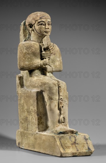 Figurine of the god Ptah. Figurine of the god Ptah sitting on a throne, on a base in the shape of the sign of Maat. He holds a composite scepter in his hands. Materials: not stated. Iconography: ankh, uas, djed pillars, back pillars, hieroglyphs, scepters, thrones. Style/period: c. 1550–c. 1295 BC; New Kingdom, 18th Dynasty, Egypt. Subject: Egyptian deity Ptah. Maker: unknown. Measurements: height 29 cm; width 10.5 cm; depth 19 cm; weight 2.736 kg., A seated stone figure on a block-like throne shown in three-quarter view, depicted with a stylized wig or hair pulled back, a defined face with carved brows and eyes, and a small beard; the figure holds a vertical object with both hands at chest level and a shorter hanging element extends downward near the knees; the surface shows a weathered patina with colors including beige, tan, light brown, gray, and small areas of blue-green corrosion; the base and throne display worn edges and textured surface variations, and the background is a smoo...