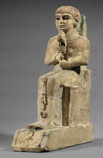 Figurine of the god Ptah. Figurine of the god Ptah sitting on a throne, on a base in the shape of the sign of Maat. He holds a composite scepter in his hands. Materials: figurine with back pillars, hieroglyphs, scepters, djed pillars, ankh and uas iconography. Style/Period: c. 1550–c. 1295 BC; New Kingdom, 18th Dynasty, Egypt. Maker: unknown. Inscriptions/markings: hieroglyphs. Subject: Egyptian deity Ptah. Function: statuette/ritual object. Dimensions: height 29 cm; width 10.5 cm; depth 19 cm; weight 2.736 kg., A seated stone figure on a rectangular base, shown in profile and frontal view, with a rounded head and stylized facial features, holding a vertical object with both hands near the chest; the figure wears a fitted garment and a layered necklace, and a long element drapes down the back. The surface shows a weathered, pale beige stone with areas of darker brown patina and small patches of blue-green corrosion. The base and lower parts of the figure display worn spots and slight d...