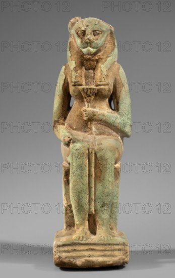 Figurine of the goddess Uadjit with the head of a lioness; Uadjit mythology; Egyptian deities; materials/details: hieroglyphs, back pillars, thrones, lotuses; maker, inscriptions: Scen.Gal.Eg., 664; date: 30 BC; period: Late Period–Ptolemaic Period; place of origin: Egypt; type: figurine; dimensions: height 22 cm, width 11.5 cm, depth 7 cm; weight 1.024 kg., A seated statue of a figure with an animal-like head wearing a striped headdress, rendered in a weathered green and tan material with areas of worn brown surface; the figure sits on a rectangular base and wears a long garment that drapes over the legs, with one arm resting across the torso and the other resting on the lap, showing textured and eroded surfaces and patches where the green finish has flaked away, set against a plain neutral gray background.