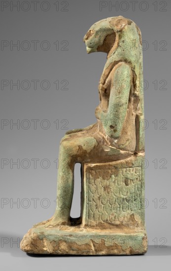 Figurine of the goddess Uadjit with the head of a lioness;, Materials/subject: Uadjit mythology, lioness-headed goddess, lotuses, hieroglyphs, back pillars, thrones. Inscriptions: Scen.Gal.Eg., 664. Date: 30 BC; Late Period - Ptolemaic Period, Egypt. Type: figurine. Dimensions: height 22 cm, width 11.5 cm, depth 7 cm; weight 1.024 kg., A seated statuette facing to the right, rendered in a weathered material showing a mix of green and tan tones, with a rounded head and a beak-like facial profile, a long smooth headdress or head covering that extends down the back, a torso with simplified arms folded or resting at the waist, a straight leg bent at the knee leading to a rectangular seat with textured surface and an open vertical slot beneath the seat, and a rectangular base that shows irregular worn edges and patches of green and tan coloration.