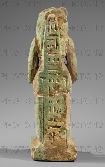 Figurine of the goddess Uadjit with the head of a lioness; Uadjit mythology; Egyptian deities; made of material not specified; hieroglyphs; back pillars; throne; lotuses; inscription Scen.Gal.Eg., 664; date: 30 BC – Late Period to Ptolemaic Period; origin: Egypt; figurine; height 22 cm, width 11.5 cm, depth 7 cm, weight 1.024 kg; maker, attributed period ca. 30 BC., A small carved figurine photographed from the back, showing a standing anthropoid form on a rectangular base; the surface is worn and pitted with a mottled green and brown coloration, and areas of lighter beige where material has flaked away; the figure has two rounded shoulder/arm forms projecting at its sides and a central vertical panel of shallow incised shapes; the base is squared with irregular edges and the overall object shows signs of age with rough texture and patches of discoloration against a neutral gray background.