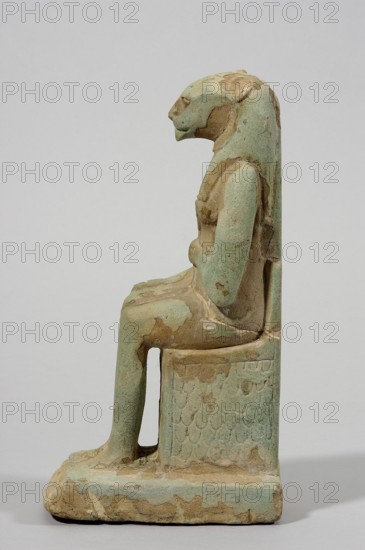 Figurine of the goddess Uadjit with the head of a lioness; maker, Uadjit mythology; depiction includes lotuses, hieroglyphs, back pillars, and throne; inscription or inventory: Scen.Gal.Eg., 664; date: 30 BC; Period: Late Period - Ptolemaic Period, Egypt; object type: figurine; dimensions: height 22 cm, width 11.5 cm, depth 7 cm; weight 1.024 kg., A side view of a small sculptural figure seated on a rectangular block, rendered in a weathered pale green with areas of tan and brown where the surface has worn away; the figure has an elongated head and a smooth, stylized profile, with a long backpiece extending down behind the shoulders and a visible arm resting close to the torso; the seated posture shows one leg bent at the knee with a hollow space beneath the thigh and a foot resting on a textured base that echoes the pale green and tan coloration, while the block beneath the figure displays a repeating scale-like pattern and mottled patches of exposed brown.