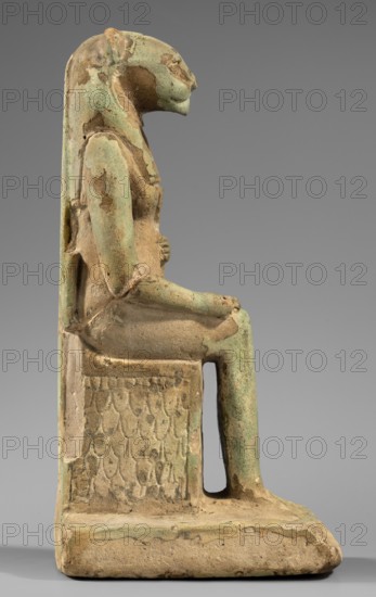 Figurine of the goddess Uadjit with the head of a lioness; Uadjit mythology; Egyptian deities; hieroglyphs; back pillars; thrones; lotuses; maker, dated 664–30 BC, Late Period–Ptolemaic Period, Egypt; figurine; height 22 cm, width 11.5 cm, depth 7 cm, weight 1.024 kg., A seated figure sculpture shown in profile with a birdlike head, elongated headdress, and a body rendered with worn surfaces and patches of green and tan; the figure sits on a rectangular throne base that has a textured panel on its side and shows areas of erosion and roughness, with the overall colors visible being green, tan, and beige against a neutral gray background.